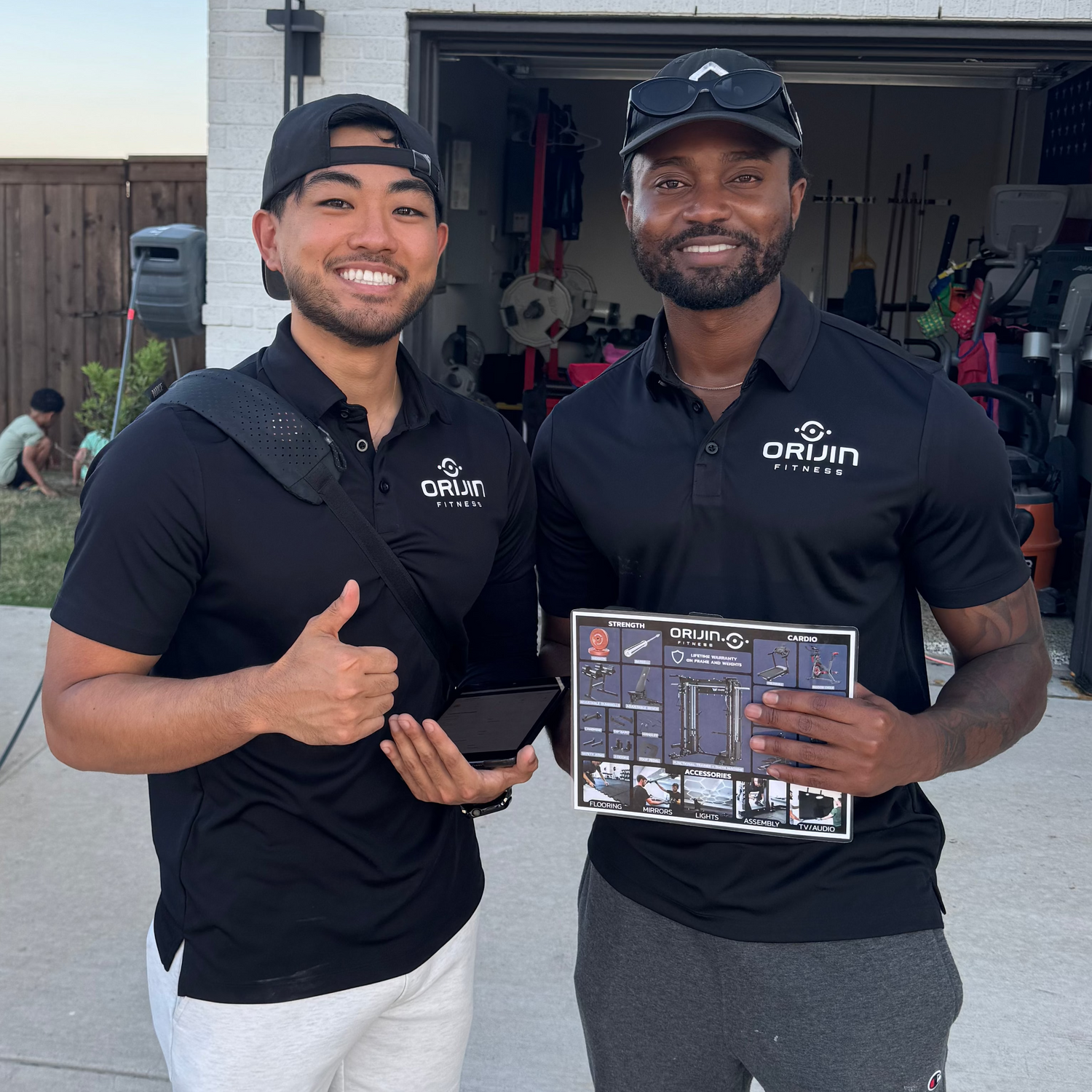 Two men in matching black shirts with logos standing outside a garage.